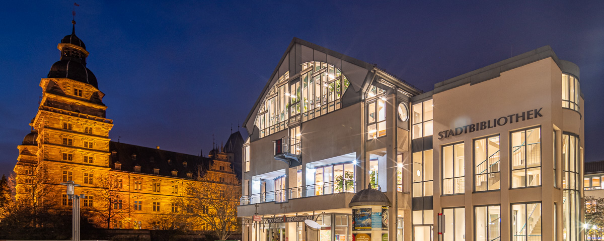 Aufnahme der beleuchteten Stadtbibliothek bei Nacht, im Hintergrund sieht man das Aschaffenburger Schloss.