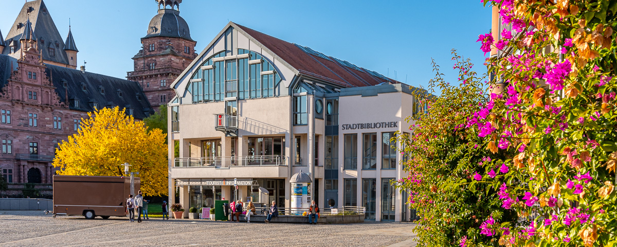 Außenansicht der Stadtbibliothek vom Marktplatz aus, an der rechten Seite sind lila blühende Blumen zu sehen, auf der linken Seite ein gelber Herbstbaum