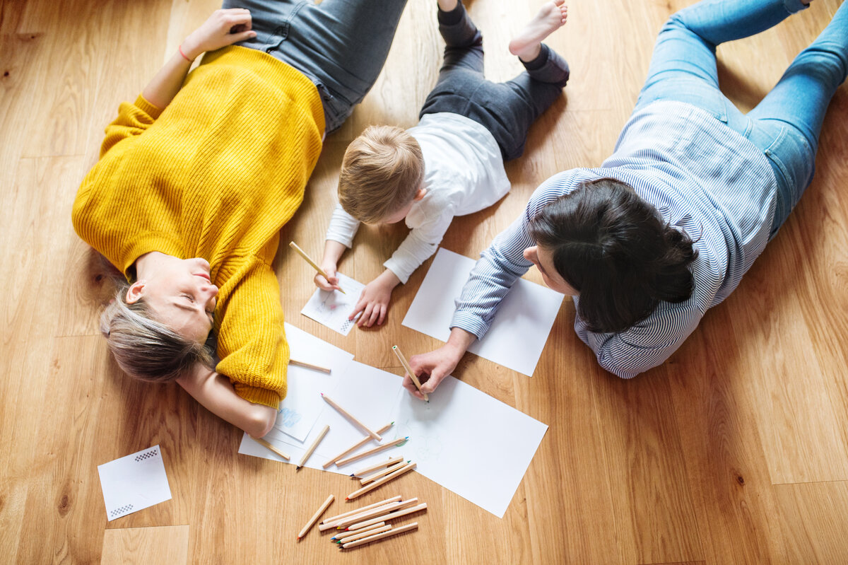 Vogelperspektive auf eine Familie liegend auf dem Fußboden, links und rechts je eine erwachsene Person, ein Junge in der Mitte, der ein Bild malt, es liegen Papier und Holzstifte zwischen den beiden Erwachsene