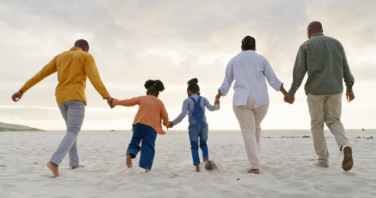 Man sieht von hinten drei Erwachsene und zwei Kinder, die sich an den Händen halten und am Strand entlanglaufen
