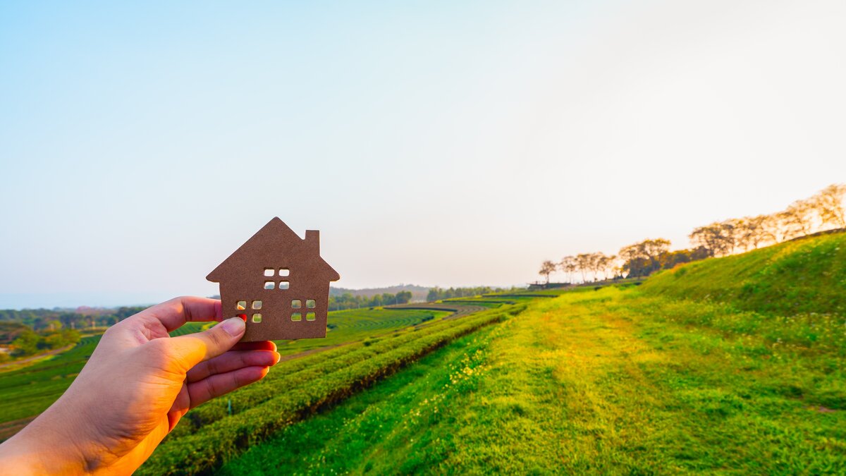 Eine Landschaft, auf der Häuser gebaut werden könnten. Im Vordergrund hält eine Hand ein Modellhaus.  