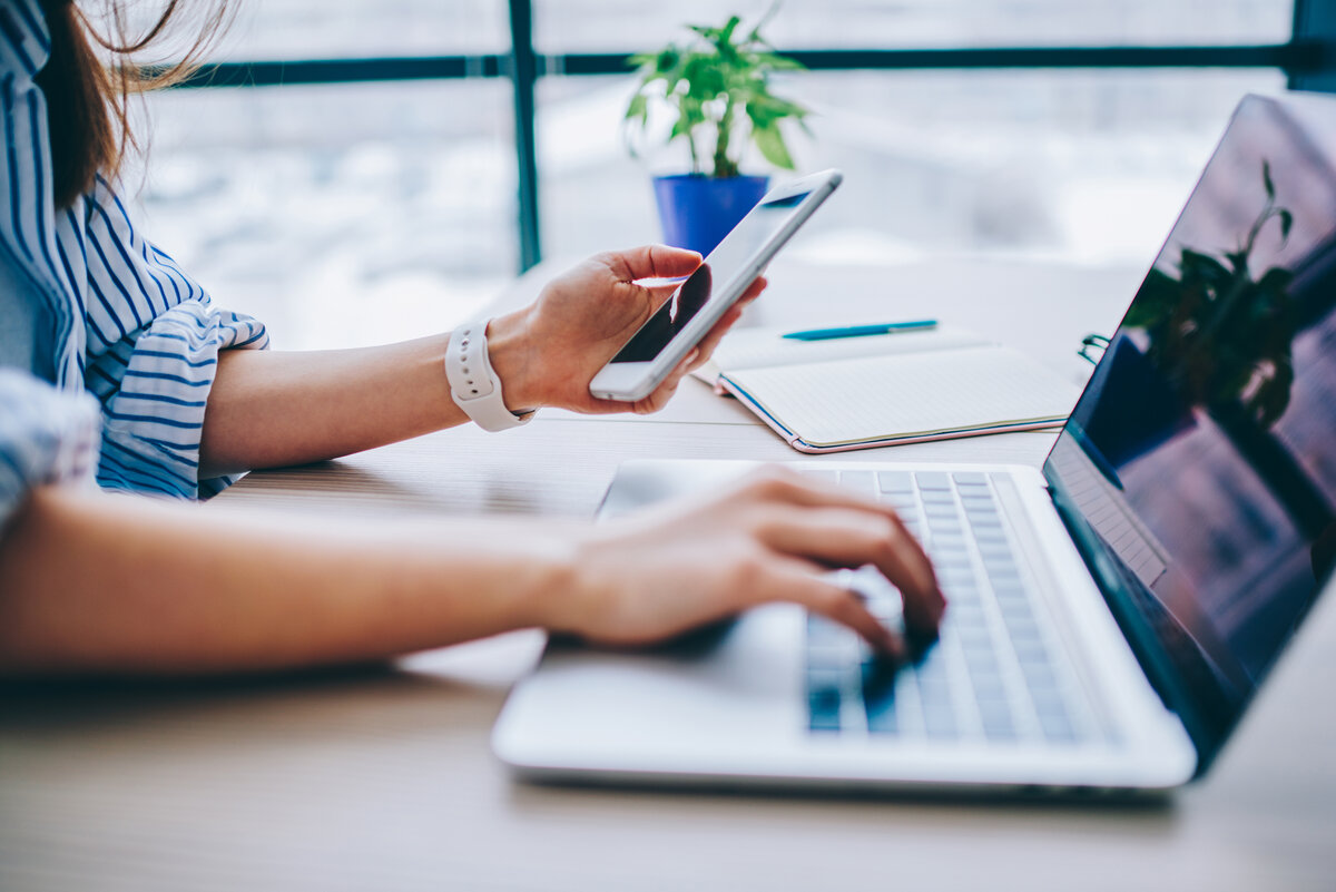 Eine Frau arbeitet am Schreibtisch mit einer Hand an einem Laptop. In der anderen Hand hält sie ein Smartphone. 