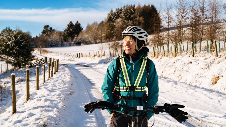 Frau mit Fahrradhelm und reflektierender Sicherheitsweste steht mit ihrem Fahrrad auf einem verschneiten Feldweg; im Hintergrund sind winterliche Bäume, Zäune und ein sonniger, blauer Himmel zu sehen.