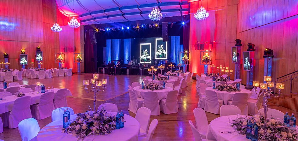 Großer Veranstaltungssaal mit heller Holzvertäfelung. Zahlreiche runde Tische sind mit weißen Tischdecken, Blumenarrangements und Kerzenleuchtern geschmückt. Im Hintergrund eine Bühne mit Vorhängen, Scheinwerfern und digitalen Leinwänden, die stilisierte