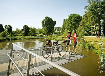 Auf einem kleinen Vorsprung über einem See stehen zwei Radelnde und genießen die Sonne, im Hintergrund ein Park